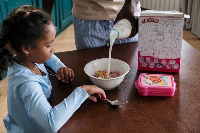 A man pouring milk into a cereal while a girl is watching. The cereal box is on the table which can be used as an eco-friendly gift packaging.